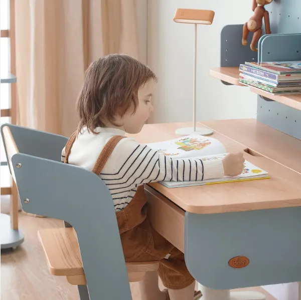 A young child sitting at a Boori natural wood and dusty blue ergonomic desk, reading a book. The desk features a built-in shelf with books and a modern desk lamp.