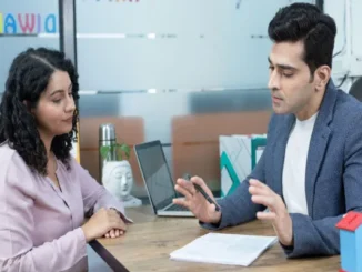 A professional insurance advisor in a blazer gestures while explaining policy details to a woman seated across a desk in a modern office.