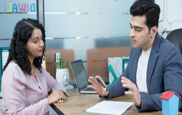A professional insurance advisor in a blazer gestures while explaining policy details to a woman seated across a desk in a modern office.