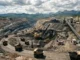 A wide panoramic view of an open-pit coal mine featuring heavy machinery, excavators, and haul trucks transporting coal against a backdrop of mountains.