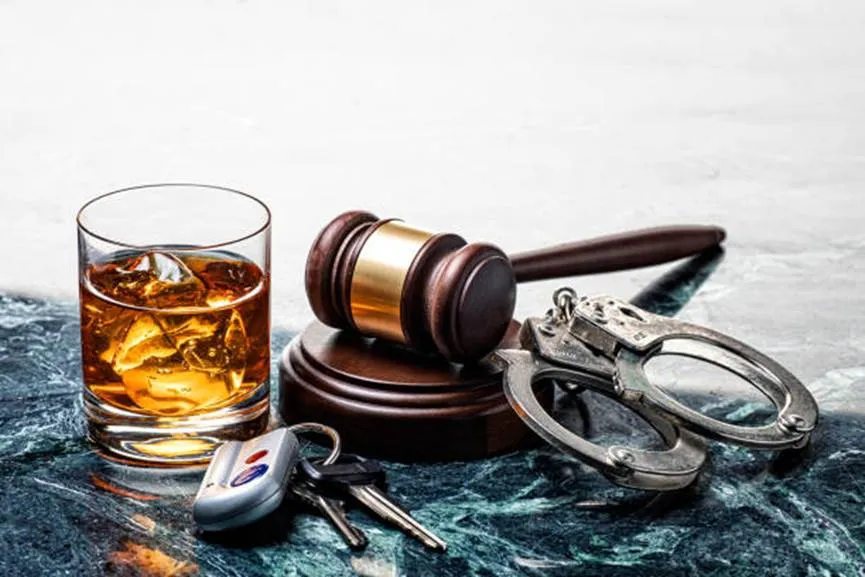 A glass of whiskey with ice sitting next to a set of car keys, a judge's wooden gavel, and a pair of silver handcuffs on a dark marble surface.