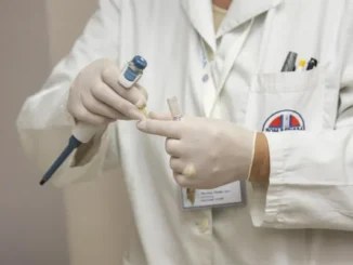 A close-up of a laboratory technician in a white coat and gloves using a precision pipette to transfer a liquid sample into a test tube for analysis.