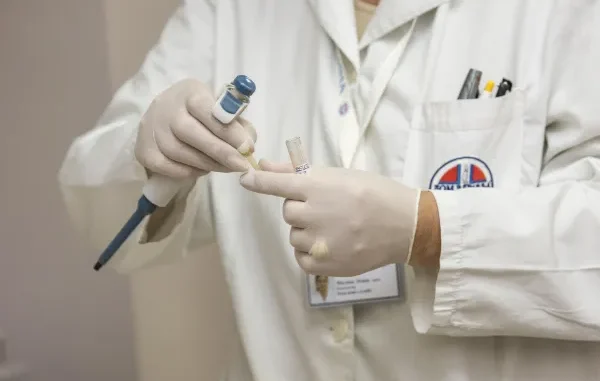 A close-up of a laboratory technician in a white coat and gloves using a precision pipette to transfer a liquid sample into a test tube for analysis.