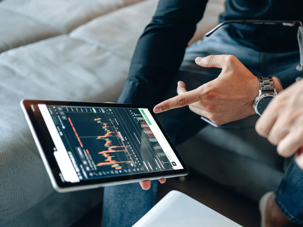 A close-up of a student pointing at a digital tablet displaying real-time stock market candlestick charts and financial data analytics.