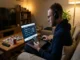 A man with headphones sitting on a sofa looking at the Jalalive TV football match schedule on his laptop while checking a sports betting app on his smartphone.