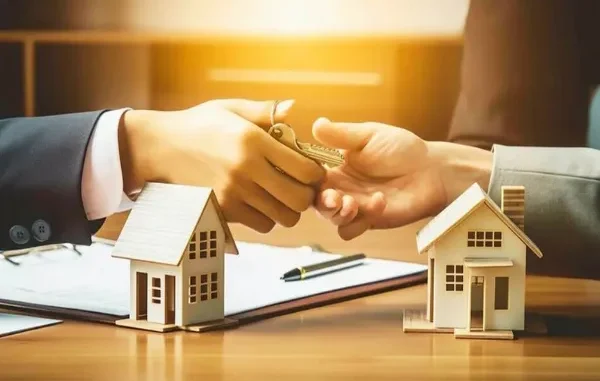 A close-up of a business owner receiving property keys over a desk with miniature house models and loan documents, symbolizing a loan against property.