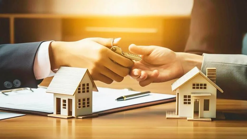 Close-up of a real estate agent handing over house keys to a business owner over a wooden desk featuring miniature model houses, a pen, and loan documents, symbolizing property-backed financing.