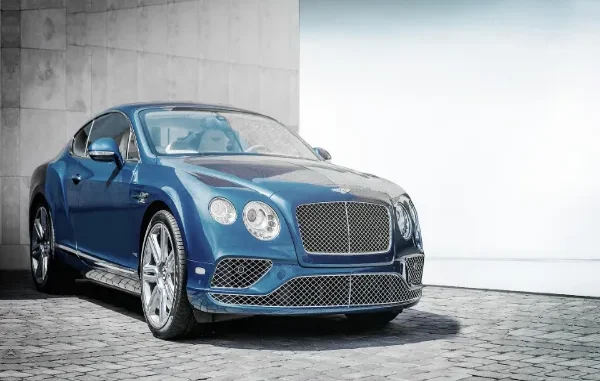 A luxury blue Bentley Continental GT parked on a stone driveway in front of a modern building, representing premium chauffeur services in Amsterdam.