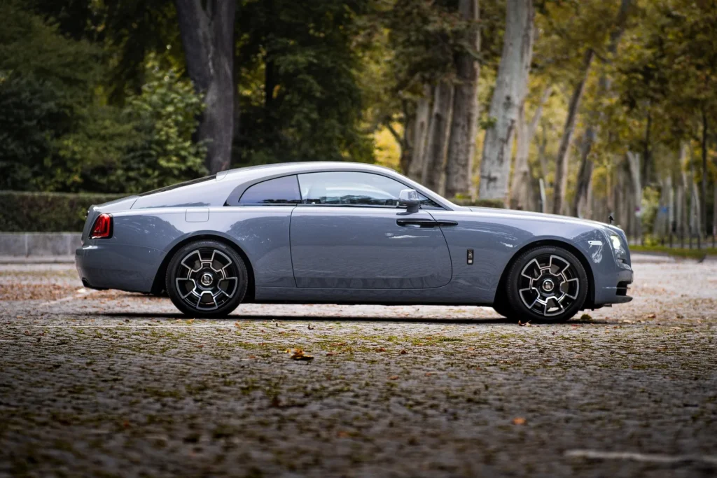 A side profile of a sleek grey Rolls-Royce Wraith parked on a cobblestone path lined with trees, representing luxury chauffeur options in Amsterdam.
