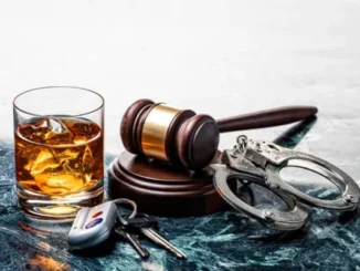 A glass of whiskey with ice, car keys, a wooden judge’s gavel, and metal handcuffs arranged together on a dark marble surface, symbolizing the legal consequences of drunk driving.