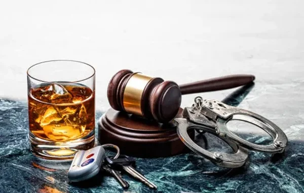 A glass of whiskey with ice, car keys, a wooden judge’s gavel, and metal handcuffs arranged together on a dark marble surface, symbolizing the legal consequences of drunk driving.