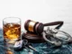 A glass of whiskey with ice, car keys, a wooden judge’s gavel, and metal handcuffs arranged together on a dark marble surface, symbolizing the legal consequences of drunk driving.
