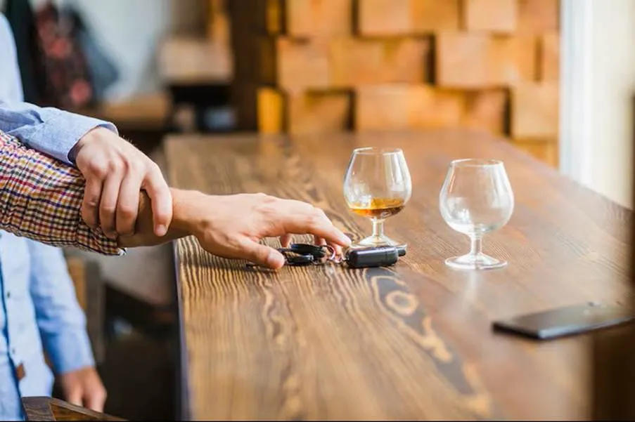 A close-up shot of one friend firmly grabbing another friend's wrist to stop them from reaching for their car keys on a wooden bar counter next to alcohol glasses.
