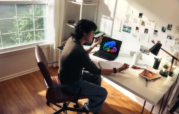 A man sitting at a sunlit white desk using a smartphone for a voice command or call while working on a laptop. The workspace is filled with physical notes and digital tools, illustrating integrated information sharing.