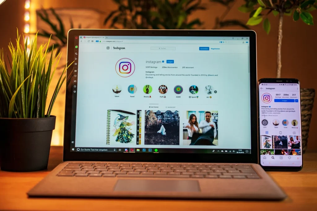 A laptop and a smartphone side-by-side on a wooden desk, both displaying the official Instagram profile page in a cozy, professional office setting with indoor plants.
