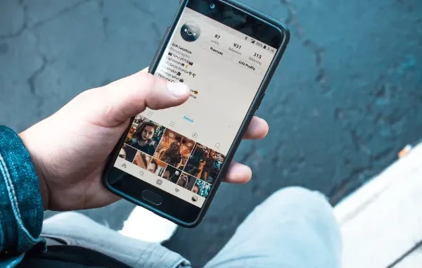 A person holding a smartphone displaying an Instagram profile page with follower counts and a photo grid, used to represent social media management and growth.