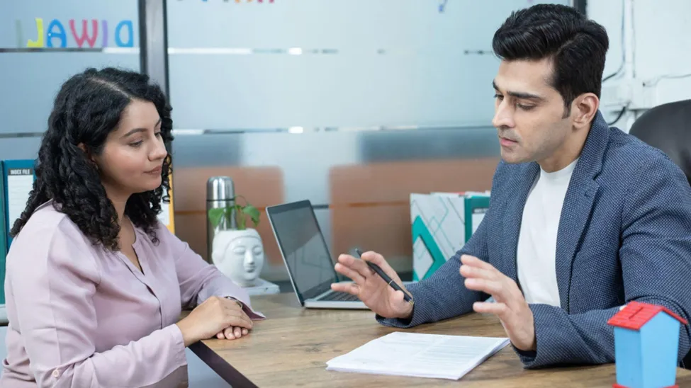 A woman listens intently as a male insurance advisor in a blue blazer explains the details of a health insurance policy document at a desk.