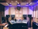 A high-end professional studio workspace featuring strategic acoustic design with wall-mounted sound panels, ceiling baffles, and ergonomic desk setup with purple LED ambient lighting.