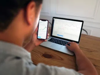 A young man sitting at a wooden desk, monitoring stock market charts on his smartphone while a laptop in the background displays a detailed trading platform.