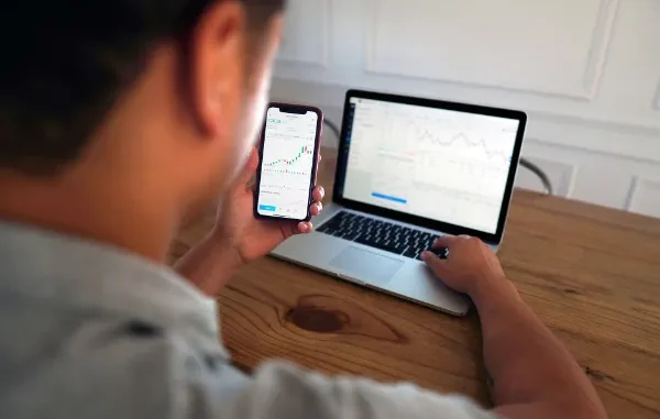 A young man sitting at a wooden desk, monitoring stock market charts on his smartphone while a laptop in the background displays a detailed trading platform.