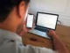 A young man sitting at a wooden desk, monitoring stock market charts on his smartphone while a laptop in the background displays a detailed trading platform.