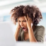 A frustrated female professional holding her head and shouting in front of a laptop, representing the stress of medical billing and lien litigation.