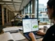 A woman with glasses sits at a library desk using a laptop that displays a split-screen view of a Plagiarism Checker (6% similarity) and an AI Content Detector (92% likely human written). Open notebooks and research papers sit on the table.
