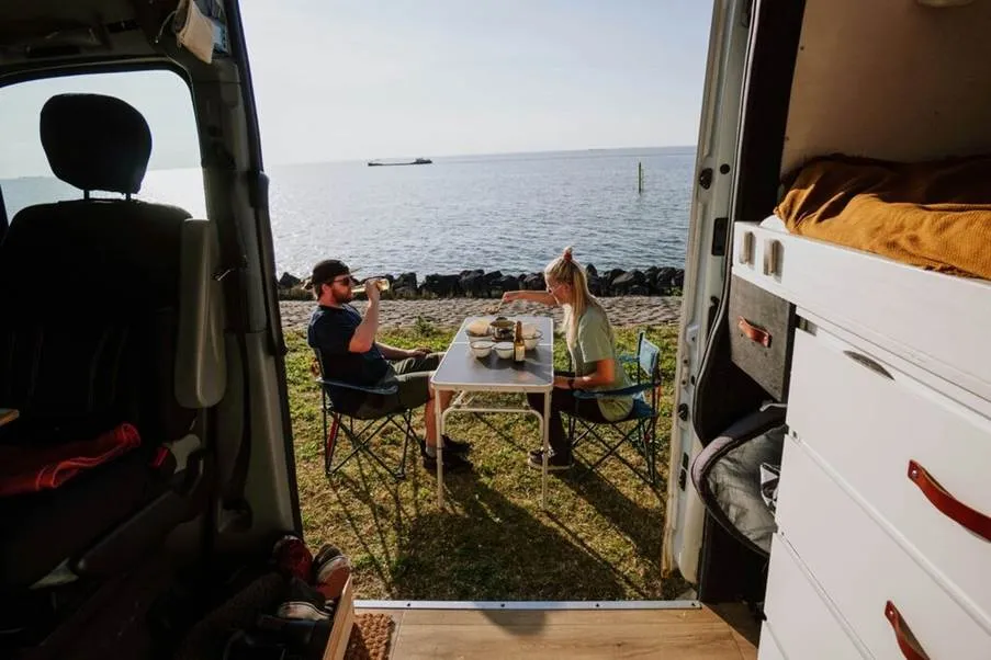 A couple enjoying a meal at a folding table next to their campervan, parked right by the ocean on a sunny day in Australia.