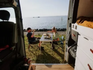 A couple enjoying a meal at a folding table next to their campervan parked by the ocean, viewed from inside the van.