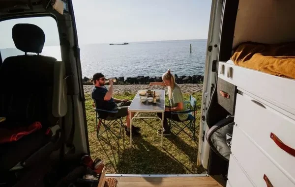 A couple enjoying a meal at a folding table next to their campervan parked by the ocean, viewed from inside the van.