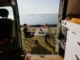 A couple enjoying a meal at a folding table next to their campervan parked by the ocean, viewed from inside the van.
