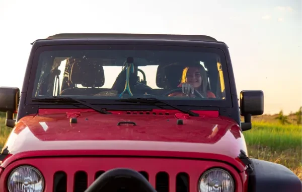 Front view of a red Jeep Gladiator with a black hardtop, featuring a smiling woman at the steering wheel in a grassy field at sunset.