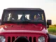 Front view of a red Jeep Gladiator with a black hardtop, featuring a smiling woman at the steering wheel in a grassy field at sunset.