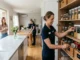 A professional home organizer from Bundl neatly arranging labeled jars and baskets inside a kitchen pantry, while another team member cleans a dining table and the homeowner relaxes in the background.
