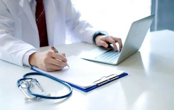 A doctor in a white coat working on a laptop and taking notes on a clipboard next to a stethoscope, evaluating locum medicine career options.