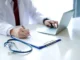 A doctor in a white coat working on a laptop and taking notes on a clipboard next to a stethoscope, evaluating locum medicine career options.