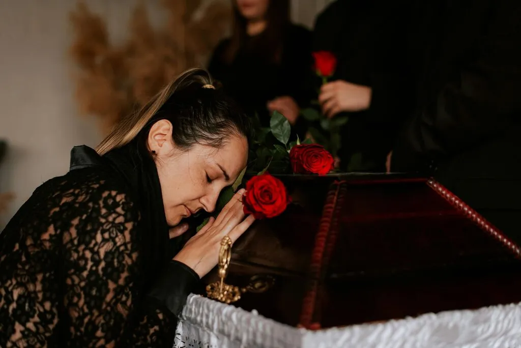 A grieving woman in black lace leans against a wooden casket with red roses during a funeral service, illustrating traditional viewing options before cremation.