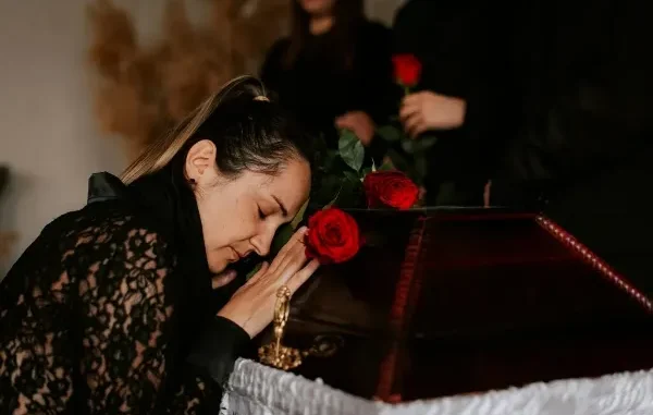 A grieving woman in black lace leaning her head against a wooden casket with red roses during a funeral service.