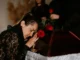 A grieving woman in black lace leaning her head against a wooden casket with red roses during a funeral service.
