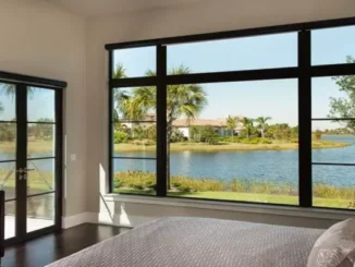 Large black-framed fixed picture windows in a modern bedroom offering an unobstructed scenic view of a lake and palm trees, paired with openable glass patio doors.