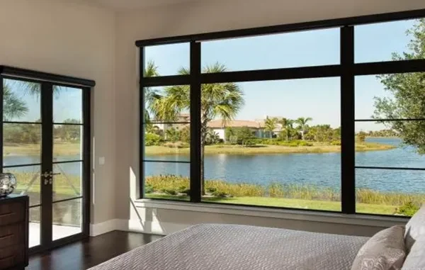 Large black-framed fixed picture windows in a modern bedroom offering an unobstructed scenic view of a lake and palm trees, paired with openable glass patio doors.