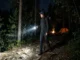 A female hiker wearing a beanie and backpack walks along a rocky forest path at night, illuminating the trail ahead with a bright handheld flashlight. An illuminated orange tent and campfire are visible in the background.
