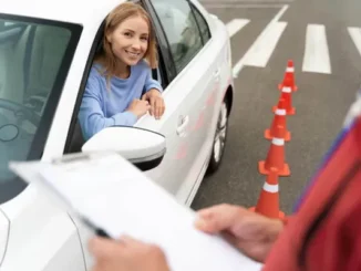 A smiling young female driver looking out of a white car window during a driving test, with an instructor holding a clipboard in the foreground and orange traffic cones in the background.