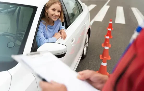 A smiling young female driver looking out of a white car window during a driving test, with an instructor holding a clipboard in the foreground and orange traffic cones in the background.