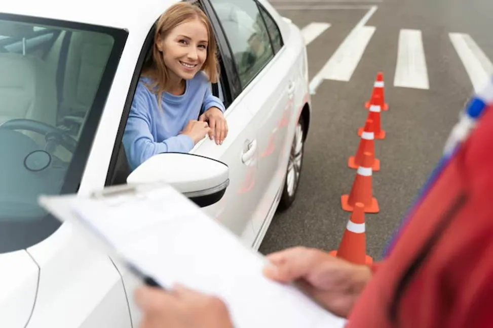 A smiling young woman sitting in the driver's seat of a white car during a driving test, looking out the window at an instructor holding a clipboard on a closed course with traffic cones.