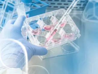A gloved hand holds a multi-well cell culture plate while a serological pipette dispenses pink liquid into one of the wells in a laboratory hood.