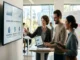 Three colleagues in a modern, open-plan office are gathered around a standing desk. The central figure, a woman in a grey blazer, points to a large wall-mounted monitor displaying charts and the title "INCREASING LIFETIME VALUE: OPERATIONAL STRATEGY." The other two colleagues, a man in a beige sweater with a laptop and a woman with a tablet, watch attentively and smile.