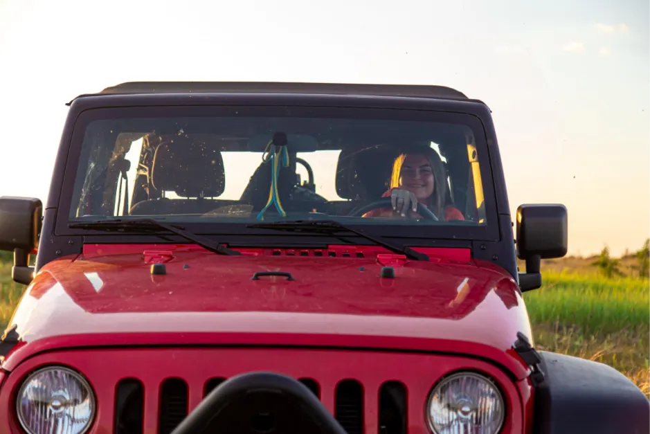 Front view of a red Jeep Gladiator with its top removed, driven by a smiling woman in a grassy field at sunset.