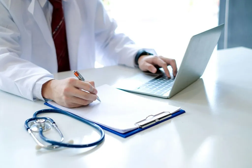 A doctor in a white coat sitting at a desk, typing on a laptop and writing notes on a clipboard with a stethoscope in the foreground, evaluating a locum tenens medical career.
