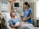 A male patient in his 40s resting in a hospital bed looks up and smiles at a supportive female nurse wearing blue scrubs. The nurse is gently checking his IV line in a bright, clean clinical room equipped with vital sign monitors, an IV stand, and a wall poster titled "Supporting Your Recovery."